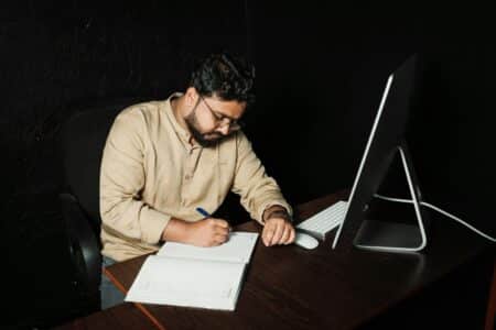 Man writing in office with computer. Late-night work focused on creativity and productivity.