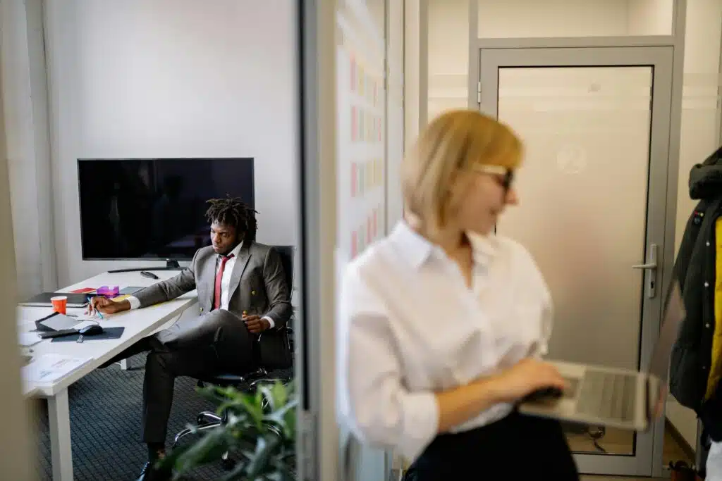 Two professionals engage in conversation within a sleek, modern office setting, one at a desk, the other with a laptop.